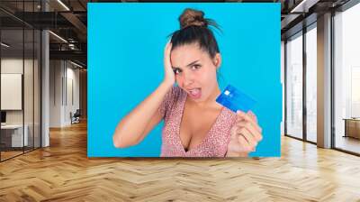 Unhappy worried beautiful brunette woman wearing floral dress over blue background keeps hand on head feels nervous holds credit card Wall mural