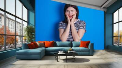 Upset young beautiful Caucasian woman wearing stripped T-shirt over blue wall touching face with two hands Wall mural