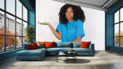 Young african woman with curly hair wearing casual blue shirt over white backgrou pointing aside with both hands showing something strange and saying: I don't know what is this. Advertisement concept. Wall mural