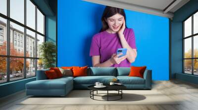 young beautiful Caucasian woman wearing purple T-shirt over blue wall holding in hands cell reading sms using new app 5g Wall mural