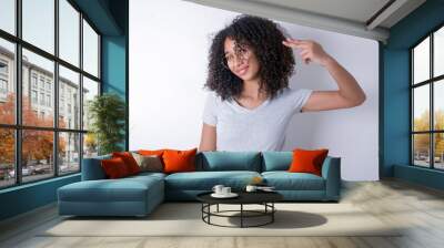 Young beautiful girl with afro hairstyle wearing gray t-shirt over white background foolishness around shoots in temple with fingers makes suicide gesture. Funny model makes finger gun pistol Wall mural