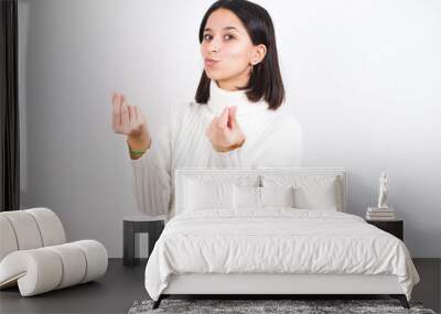 Young brunette woman wearing white knitted sweater against white background doing money gesture with hands, asking for salary payment, millionaire business Wall mural