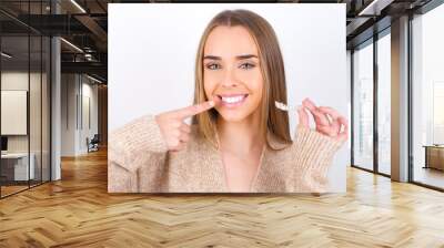 young caucasian girl wearing knitted sweater over white background holding an invisible aligner and pointing to her perfect straight teeth. Dental healthcare and confidence concept. Wall mural