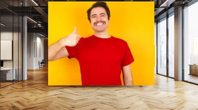 Young Caucasian man wearing red t-shirt standing against yellow background doing happy thumbs up gesture with hand. Approving expression looking at the camera showing success. Wall mural