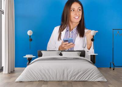 young european doctor girl wearing white medical gown ​on white background using and texting with smartphone  pointing and showing with thumb up to the side with happy face smiling Wall mural