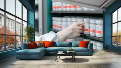 A hand in a glove reaches for a medicine bottle on a shelf, displaying a neat arrangement of pharmaceutical containers in a storage area. Wall mural