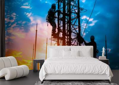 Silhouettes of two workers climbing a tall communication tower against a vibrant sunset sky. Wall mural