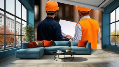 Two construction workers in hard hats review blueprints on a job site, focusing on a project under construction. Wall mural