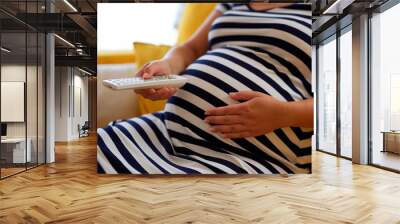 Pregnant woman sitting on sofa and holding remote control Wall mural