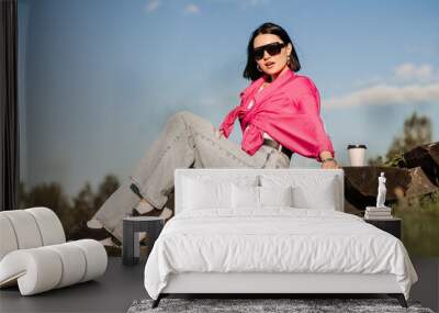 Brunette woman in sunglasses and pink shirt sitting and relaxing in the park over the blue sky background Wall mural