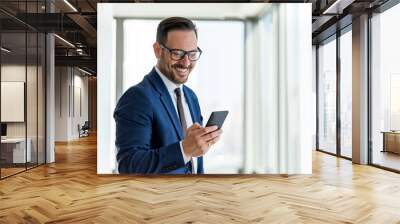 Handsome businessman wearing eyeglasses using smart phone and smiling while standing near the office window, Copy Space Wall mural