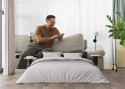 Image of a handsome cheerful happy young positive man indoors at home on sofa using mobile phone. Wall mural