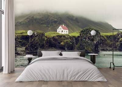 Lonely icelandic house with red roof on the sea coast with green grass meadow, rocks anf foggy sky. Natural Iceland travel landscape. Wall mural