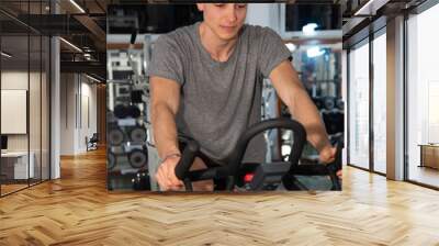 Young male is training in the gym. Use the exercise bike fitness tool similar to a bicycle and designed to reproduce the cycling movement indoors (indoors). Wall mural