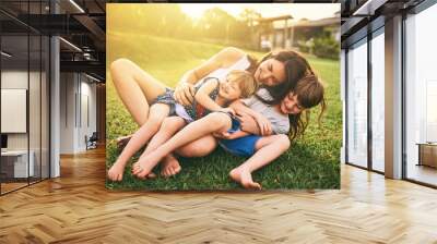 Mother, children and hug playing on grass for fun bonding in the sun outside their house in nature. Happy mom hugging kids on garden floor outdoors in playful, joy and happiness of family together Wall mural