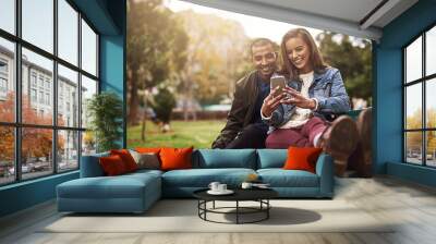 What do you think about this photo. Shot of a cheerful young couple sitting down on a bench while using a phone together outside in a park. Wall mural