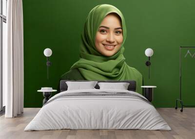 Beautiful pensive Asian girl wearing hijab smiling at empty space, isolated on green background Wall mural