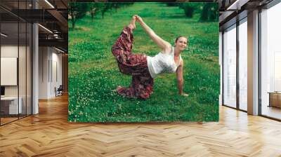 woman in a white tank top and multi-colored pants is doing yoga on the green grass in the city Wall mural