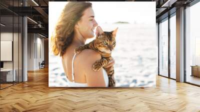 Cute girl in white dress on the beach hugs her bengal cat Wall mural