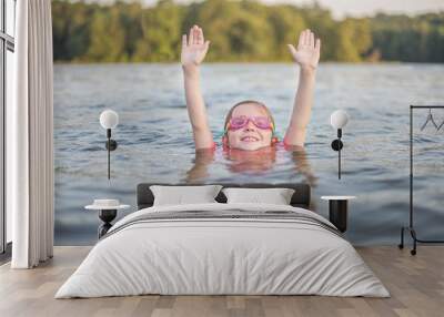 Young girl swimming in lake wearing goggles raising hands over head and poking head out of the water with forest in background and shallow depth of field Wall mural