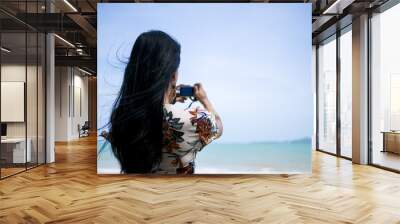 portrait of a cute asian woman wearing retro dress standing at seaside holding camera in hand and ta Wall mural