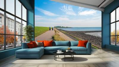 A man and a woman are cycling on a path at the bottom of a Dutch dike and along the Oosterschelde. The dike is reinforced with stones and partly covered with green algae. It is summertime now. Wall mural