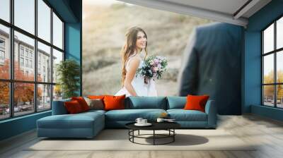 Young bride with wedding bouquet at the hands looking to groom with smile at sunset. Outdoors Wall mural