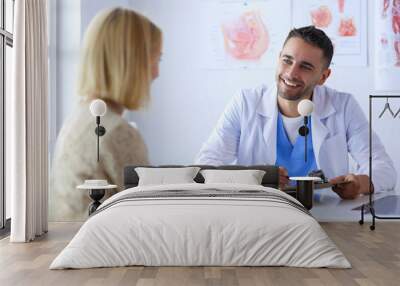 Handsome doctor is talking with young female patient and making notes while sitting in his office Wall mural