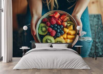 A person holds a colorful bowl filled with fresh fruits in a cozy indoor setting Wall mural