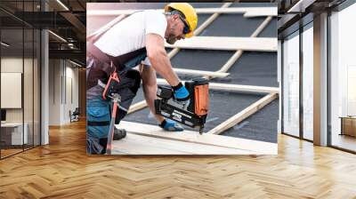 The carpenter nails a timber board using an electric nailer while working on a roof. Wall mural