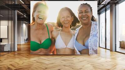 Senior multiracial women having fun hugging each other on the beach and smiling on camera during sunrise - Joyful elderly and travel vacation concept Wall mural