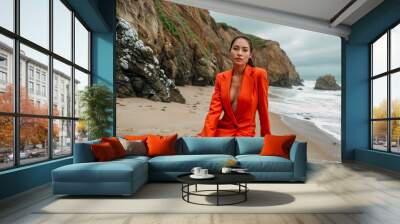 Young woman in a bright orange suit sits confidently on a black stool on a sandy beach with rocky cliffs in the background, showcasing modern beach fashion and elegance on a cloudy day Wall mural