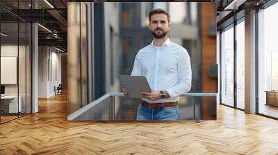 Young businessman holding laptop while standing on balcony in front of modern office building Wall mural