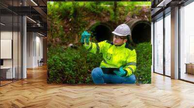 An ecologist in a green jacket sit aside of the pond looking at a liquid in a plastic glass in his hand try to examination the quality of the water. and write down the result on a clipboard Wall mural