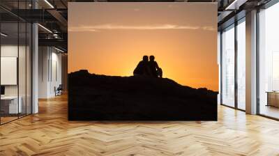 Silhouette of Couple sitting on a rock near the sea and watching the sunset Wall mural