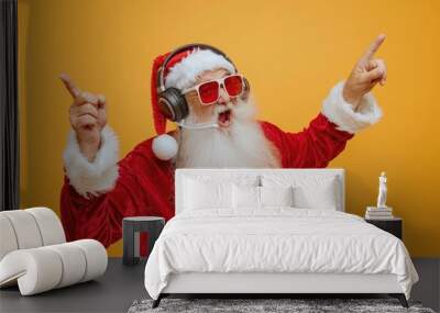 cool santa claus with headphones and sunglasses showing empty space on hands, isolated yellow color background, white gloves, funny pose, open mouth, happy face expression, red , full body portrait. Wall mural