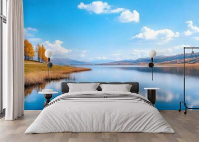 A serene landscape featuring a calm lake surrounded by mountains and vibrant trees under a beautiful blue sky. Wall mural