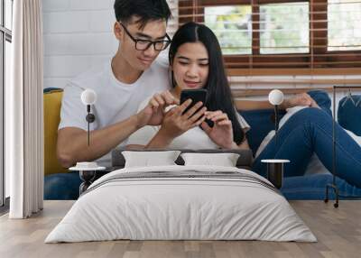 Asian young couple are looking at a smartphone in their house.Happy asian couple wearing white T-shirt and blue jeans having a good time together Wall mural