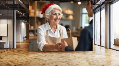 A cheerful elderly woman in a Santa hat hands a gift bag to a young person in a cozy shop during the holiday season. Wall mural