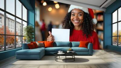 Smiling African American woman with curly hair wearing a red sweater and a Santa Claus hat,  holding a blank white card on library background Mockup.  Wall mural