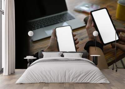 Close-up image of two hands holding a white blank screen smartphone over the working desk surrounded by an empty screen computer laptop and various stuff. Wall mural