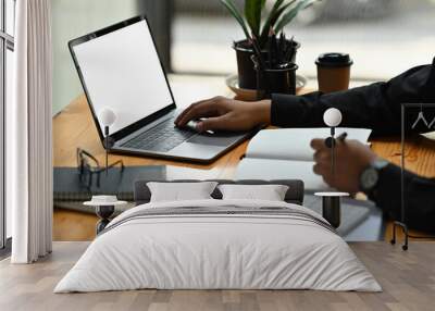 Cropped image of a young office man writing on a notebook while typing on a white blank screen computer laptop at the wooden working desk. Wall mural