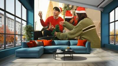 Three friends, wearing Santa hats, joyfully unpack ornaments and a Christmas tree from a storage bin, preparing to decorate their space for the holiday season, Setting Up for a Christmas Celebration Wall mural