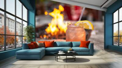 A fruity cocktail drink with a lemon slice served on a mason jar, with a table top fire pit out of focus in the background Wall mural