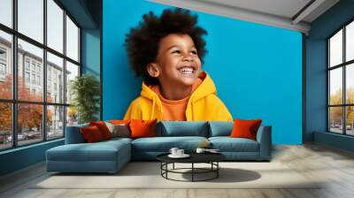 Portrait of a young black boy laughing and smiling wearing yellow hoody on bright blue background Wall mural