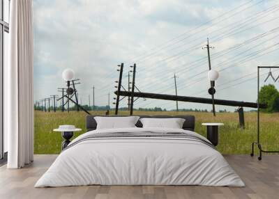 Two wooden poles of the power line broke and fell. The communication line goes into the distance. Supports stand in a blooming field. In the background, a cloudy sky, forest and concrete pillars  Wall mural