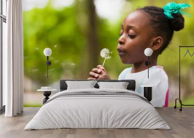 Outdoor portrait of a cute young black girl blowing a dandelion Wall mural