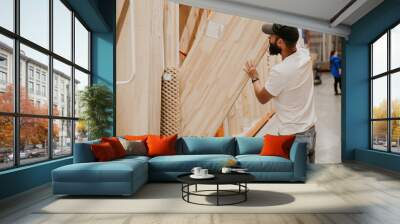 A young handsome man with a beard in casual clothes in a construction hypermarket in the lumber department selects wood building materials for the renovation of his house. Wall mural