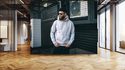 City portrait of handsome hipster guy with beard wearing gray blank hoodie or hoody and hat with space for your logo or design. Mockup for print Wall mural