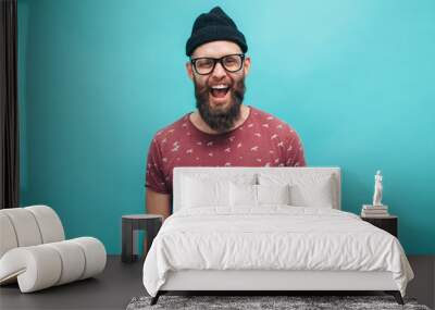 Handsome hipster guy with beard smiling happily, dressed casually with black beanie on head isolated over a blue studio background. The concept of people, crazy emotions. Wall mural